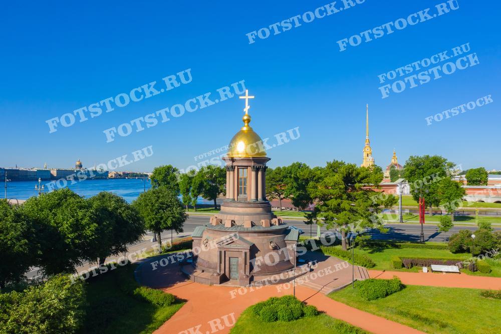 На фотографии изображена часовня-памятник на Троицкой площади в Санкт-Петербурге. Вдалеке виднеются золотые шпили и купола Петропавловской крепости, расположенные на берегу Невы. Часовня в центре снимка выделяется своими пропорциями и архитектурным стилем, подчёркивая историческое и культурное значение этого места. Это изображение может использоваться в материалах, посвящённых истории города, культурном туризме или как элемент дизайна в печатных и цифровых изданиях.